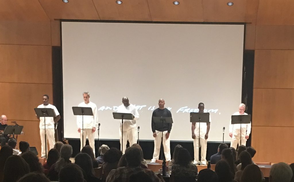 Six individuals stand on a stage in front of individual podiums. They are all dressed in black and white and are addressing a crowd in front of them during a performance of On the Row at the 2017 Conference on Community Writing.