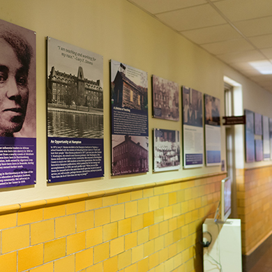 An empty hallway made of yellow brick and white walls. Informational posters about Lucy F. Simms are hanging on the left side of the hallway.