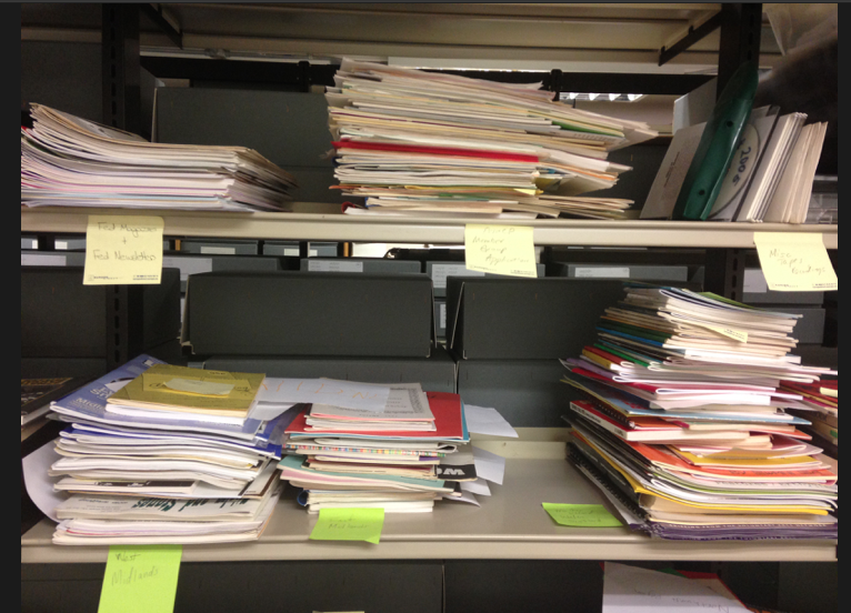 A side view of several stacks of historic magazines, books, and other forms of publications, organized on a shelf with sticky note labels.