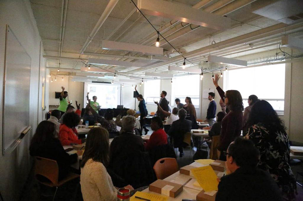 People of various genders and races sit at tables in a presentation room. Several people are standing, with their hands raised to ask questions.