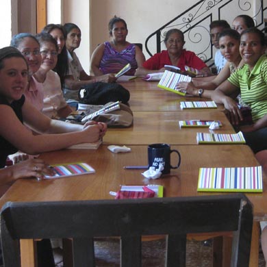 A group of women sit around a table with notebooks in front of them. Many of them are turned and face the camera, smiling.