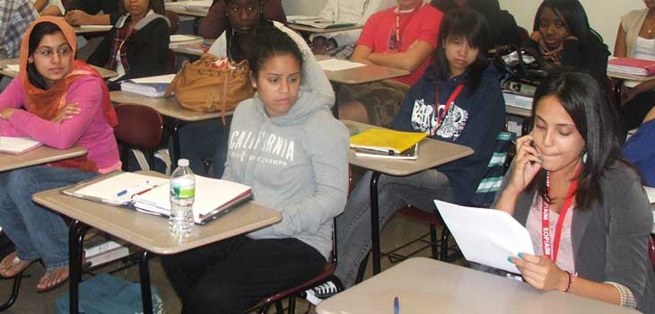 A group of teens sit at desks with open notebooks in front of them. They are looking to and listening to one of the teens as she reads aloud.