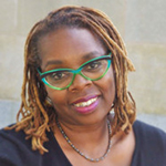 A Black woman in green glasses smiles open-mouthed at the camera.