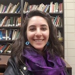 Image description: A person with dark hair stands in front of a bookshelf smiling at the camera.